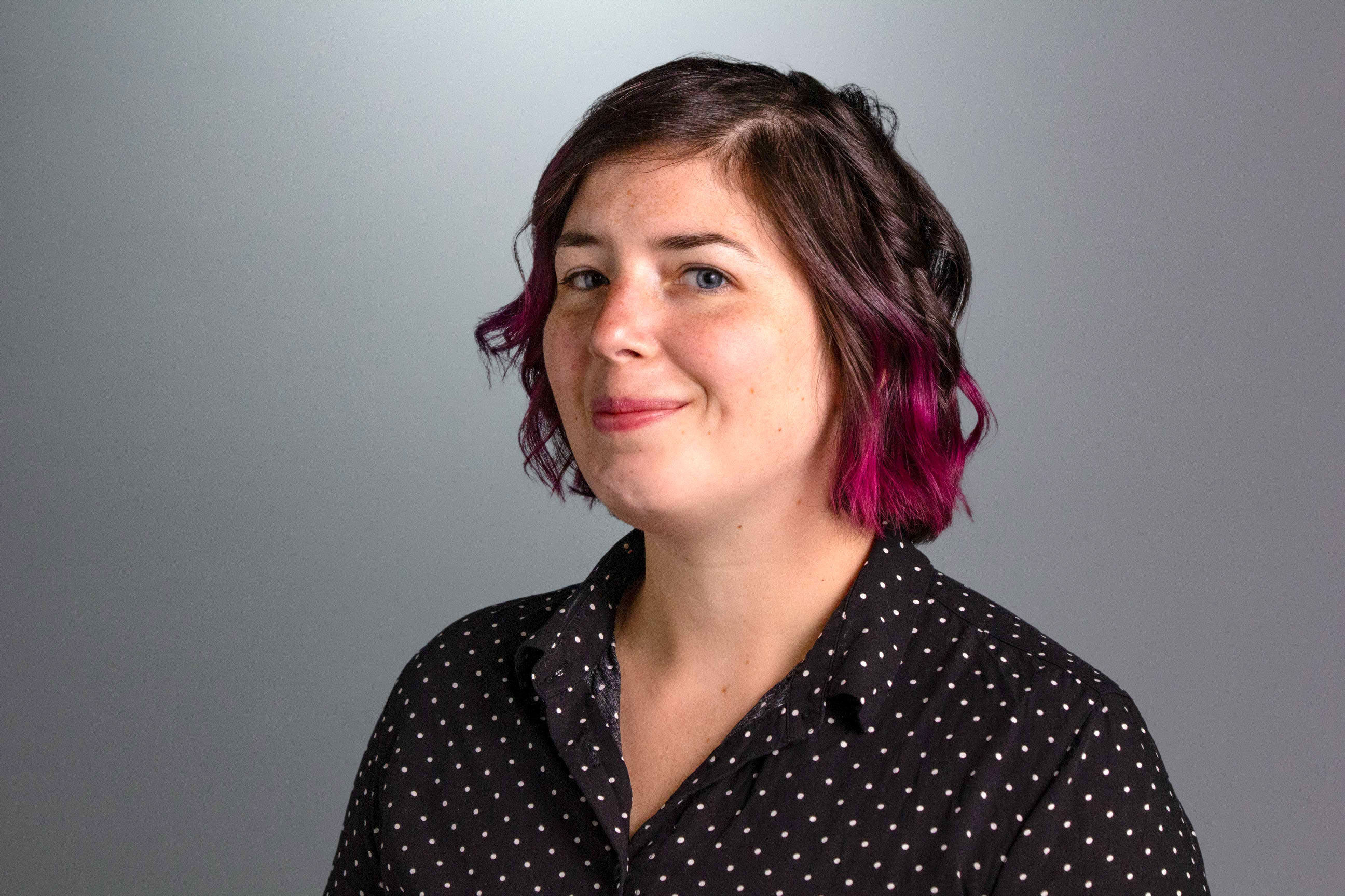 Ash headshot, she has short brown hair with pink tips and is wearing a dark button down shirt with white polka dots. 