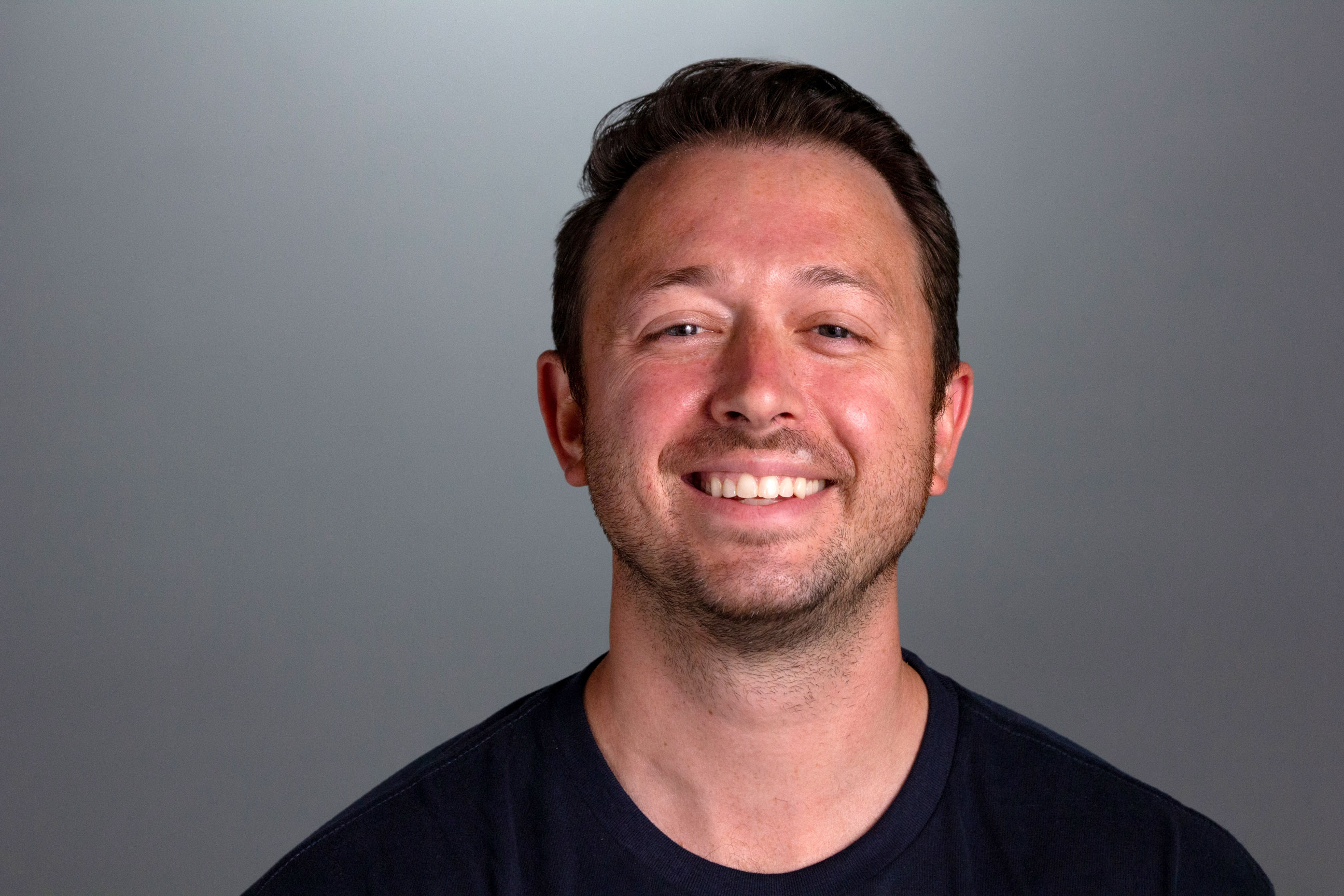 Brad sits in front of a gray background smiling at the camera wearing a dark blue tshirt
