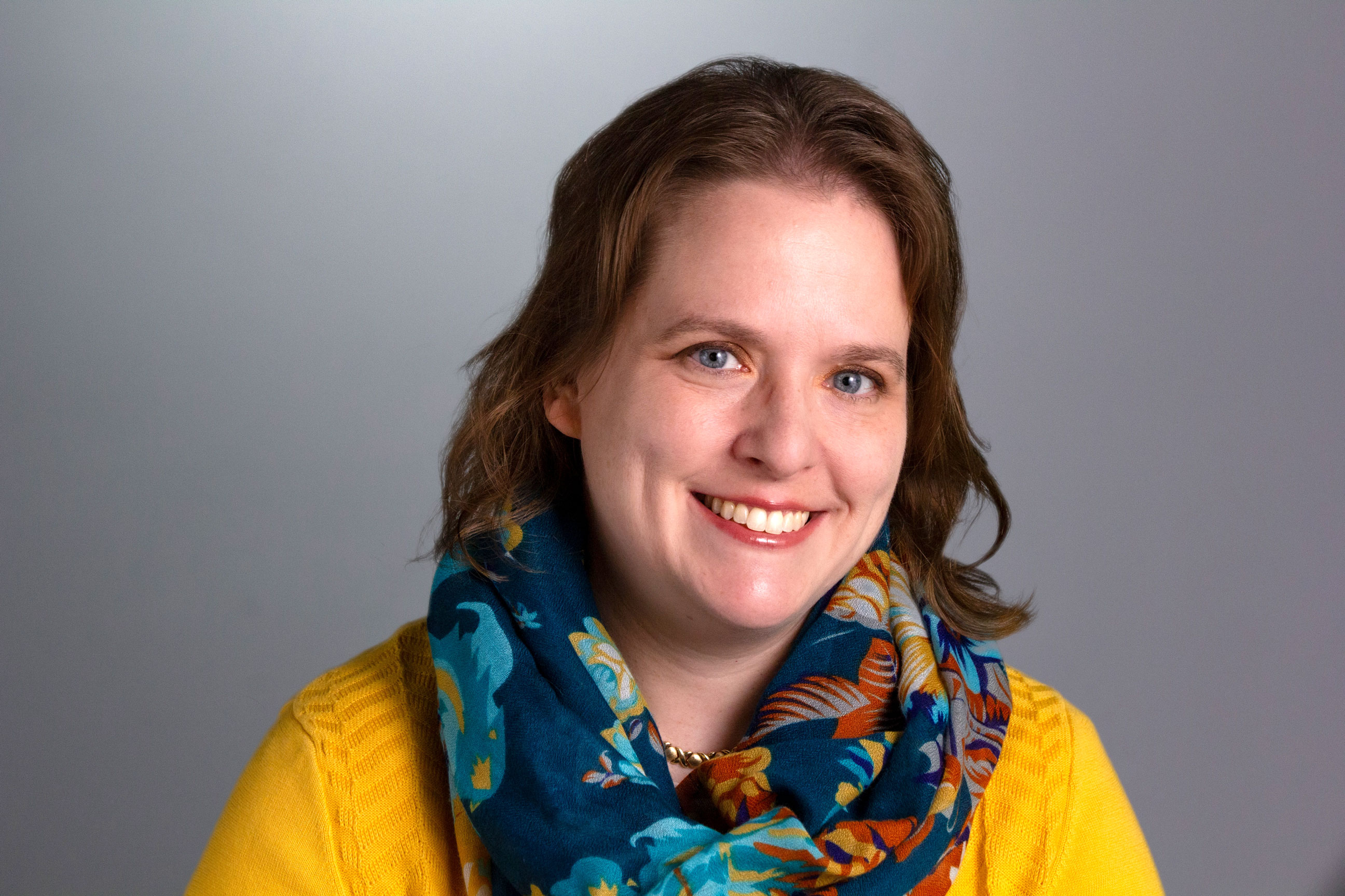 Headshot of Brenda, who is wearing a bright yellow sweater and a blue and brown patterned scarf. She has medium length dark blonde hair. 