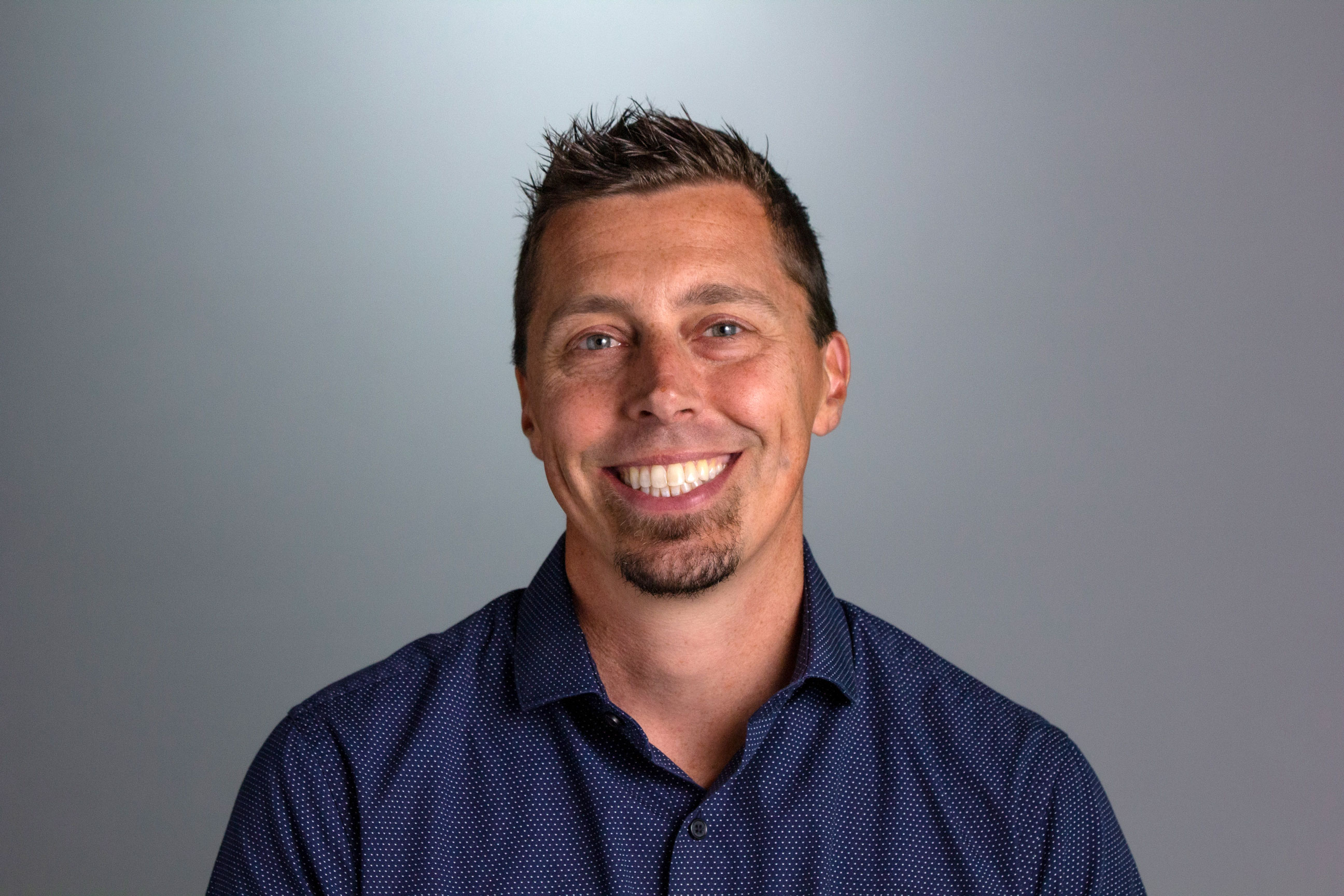 Headshot for Brendan, he is seen against a gray background and is wearing a blue collared shirt. 