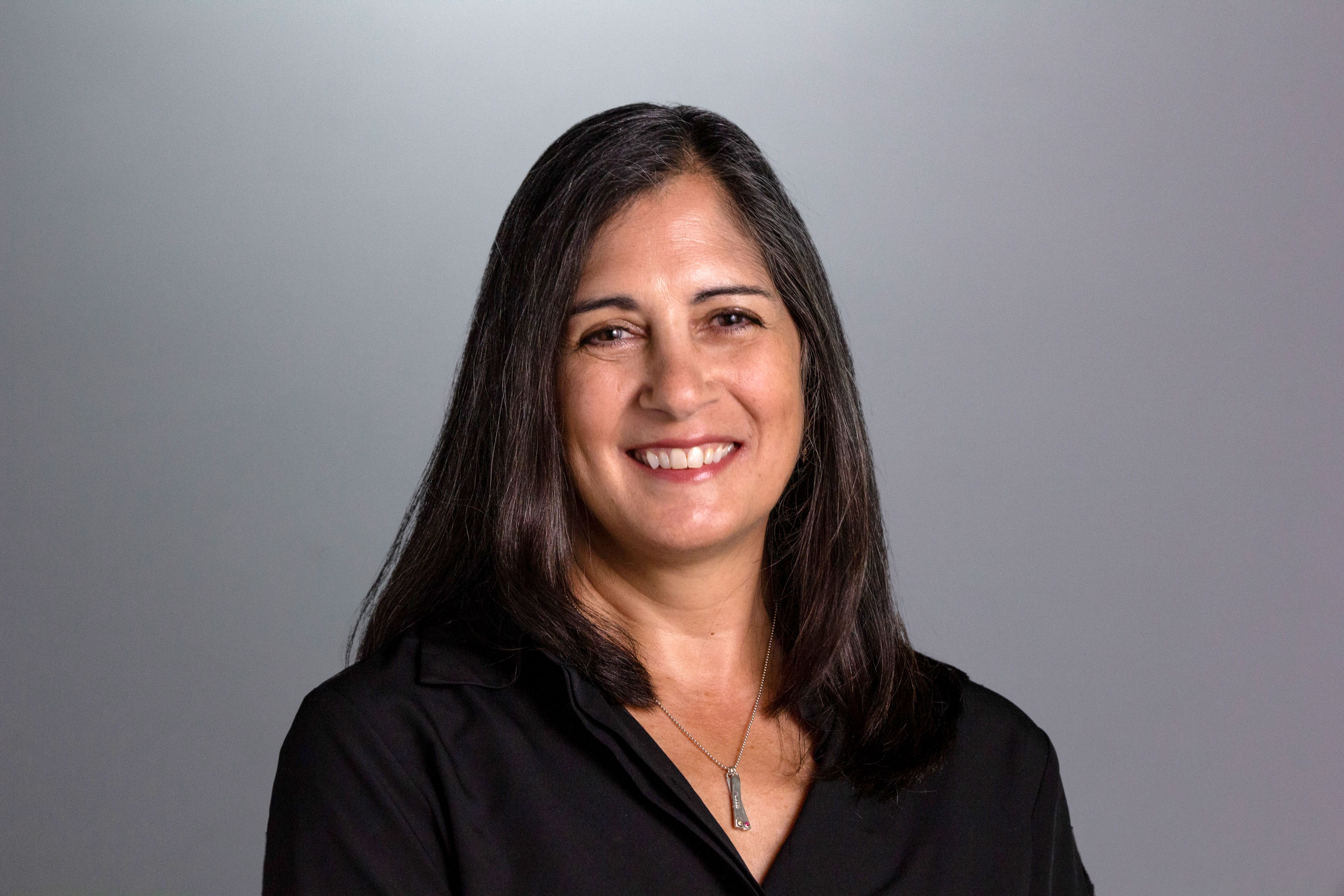 Diane sits in front of a gray background wearing a black shirt, a simple necklace, and has dark brown hair. 