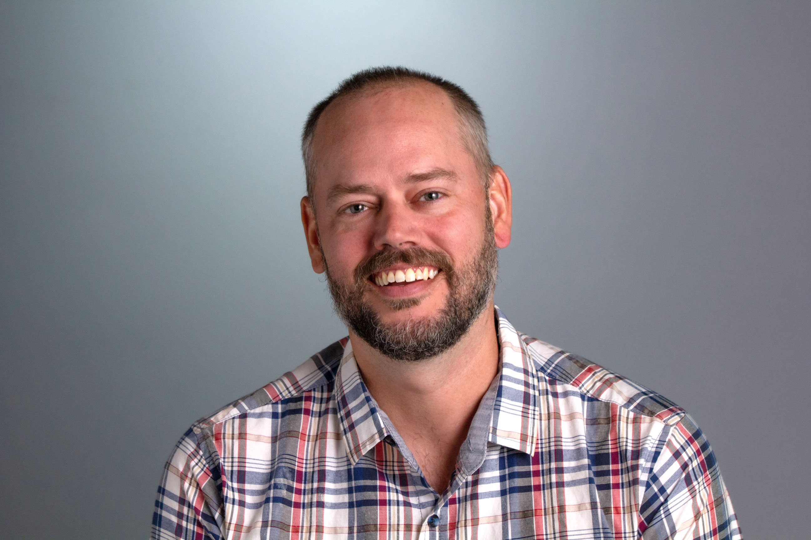 Jared sits in front of a gray backdrop wearing a multi-colored plaid button down shirt