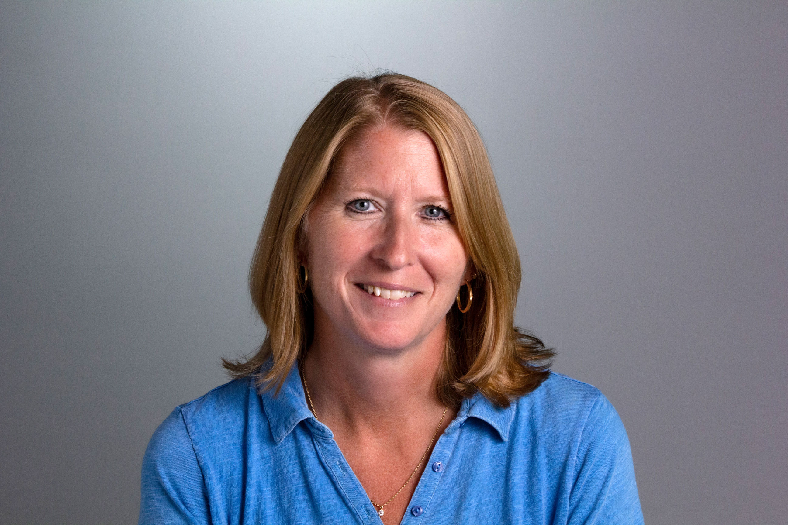 Jennifer is wearing a light blue blouse and has blonde hair and blue eyes and is in front of a gray background