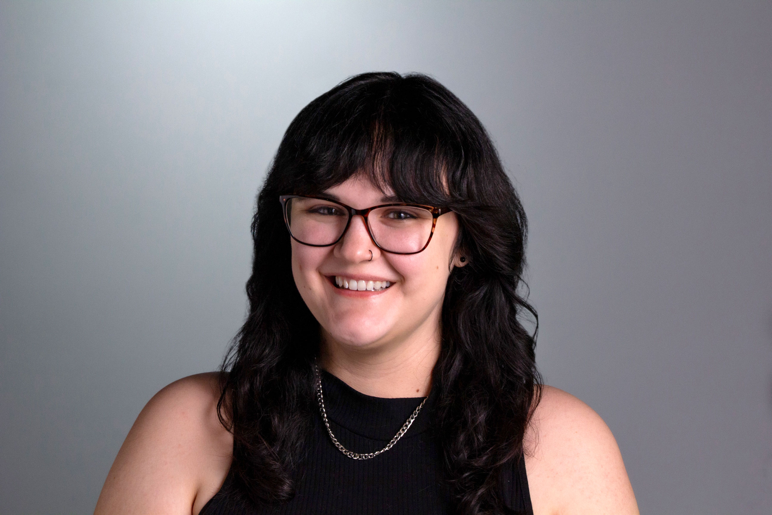 Krystal sits in front of a gray background in a black tank top with medium-long dark hair in soft waves with bangs