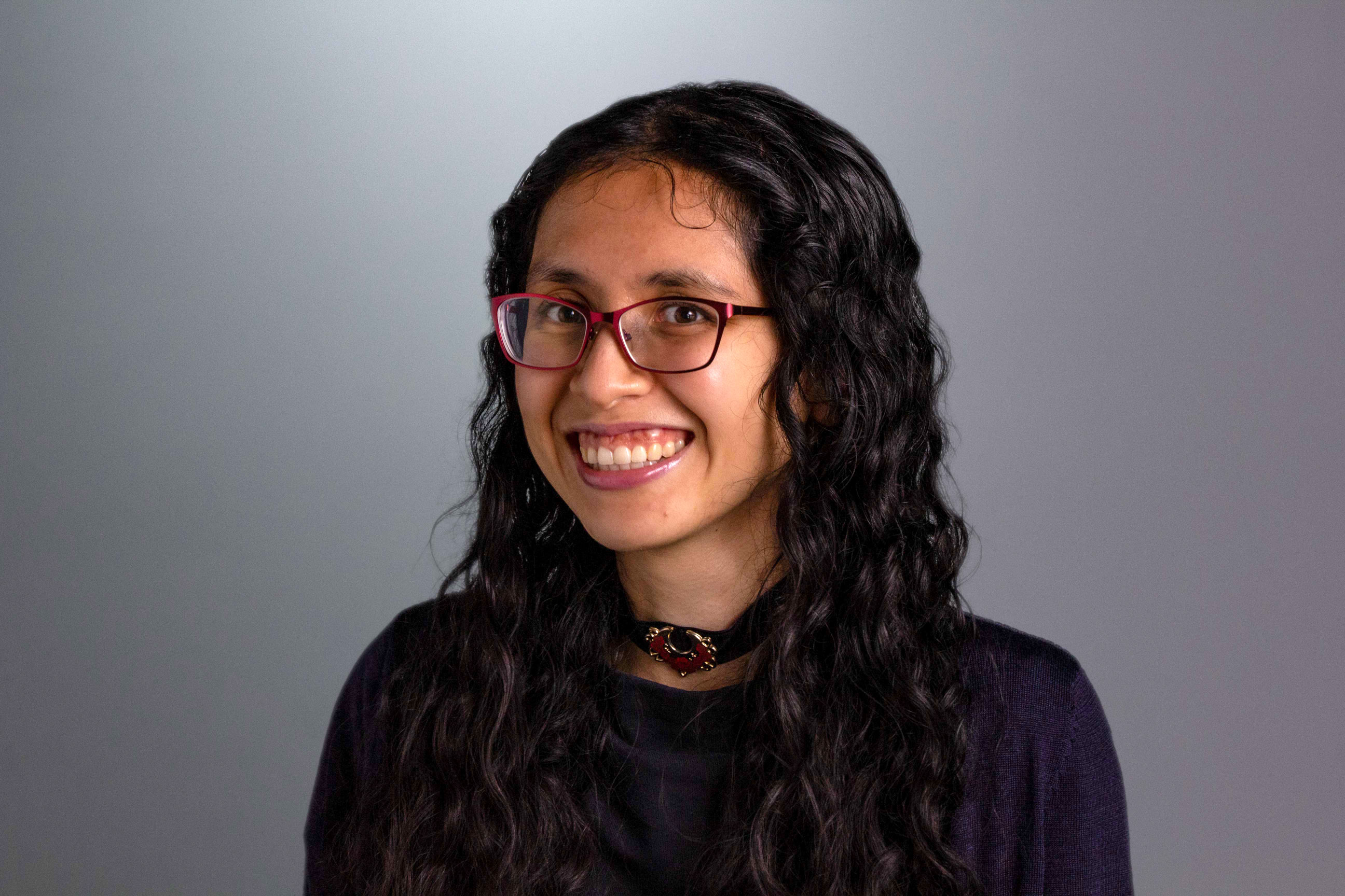 Lauren is seen in front of a gray backdrop with dark wavy hair wearing a chunky necklace and a plum colored shirt