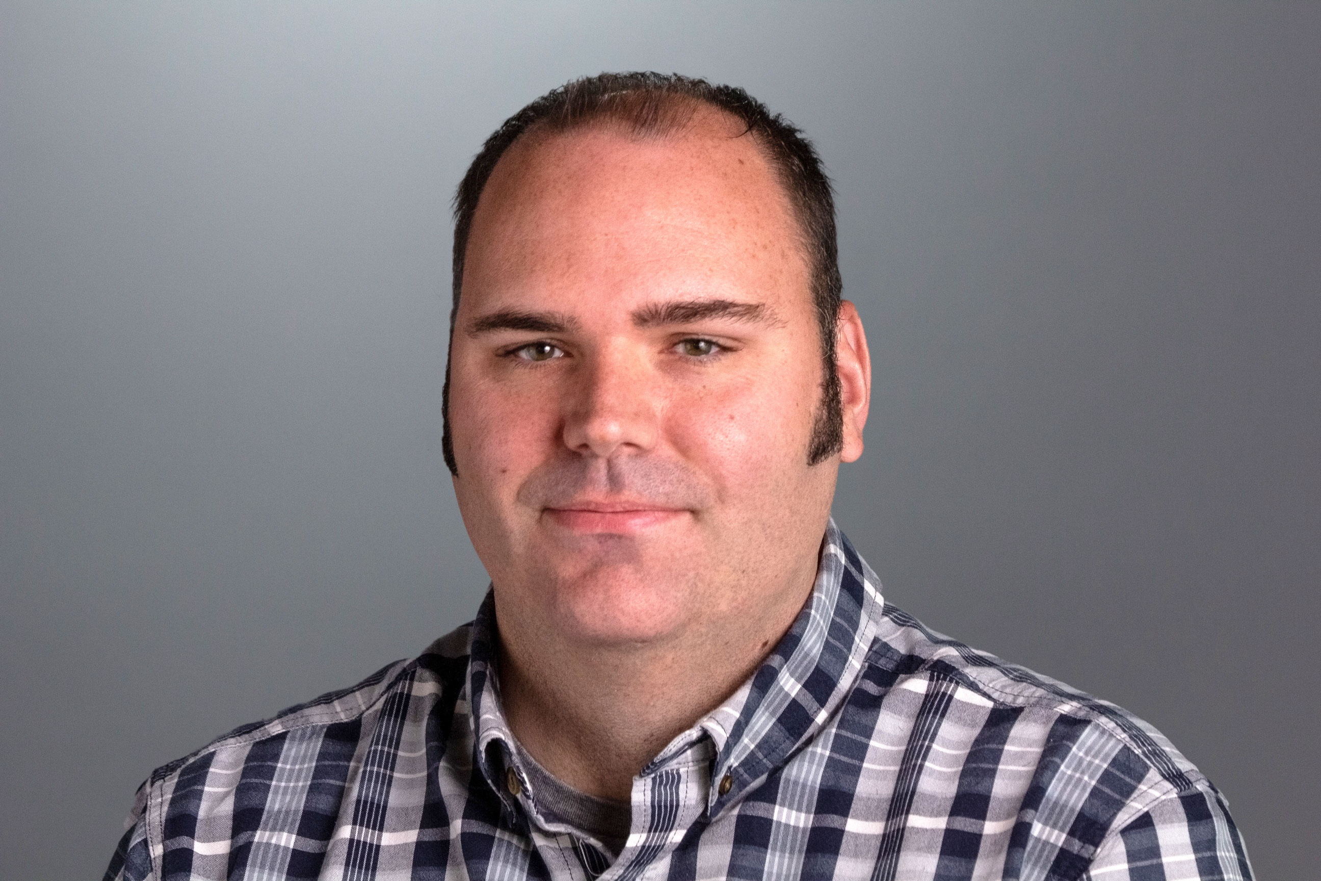 Matt sits in front of a gray background and is wearing a black and gray plaid button down shirt. 