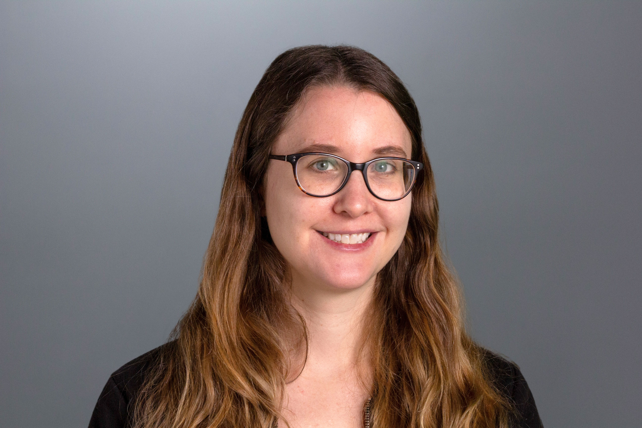 Megan sits in front of a gray backdrop wearing a dark colored shirt, glasses, and long blonde hair