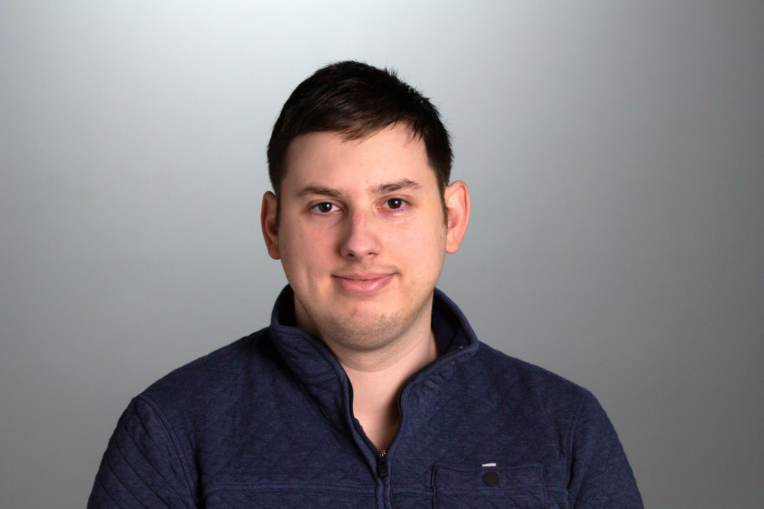 Headshot for Mike Adams, against a gray background and wearing a dark blue half zip jacket.