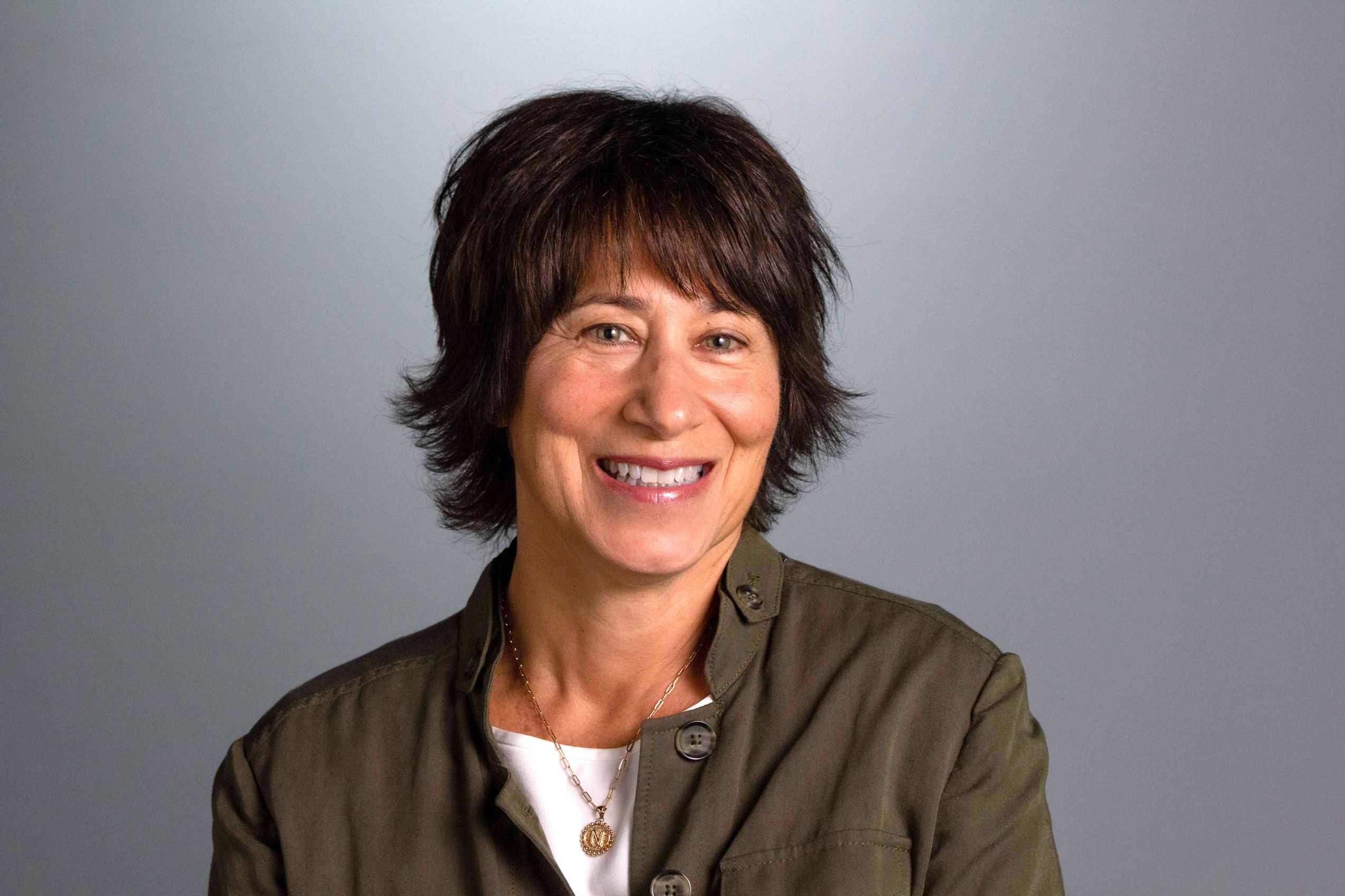 Nancy has short brown hair and is wearing a forest green outer layer with a white shirt underneath. She is in front of a gray background. 