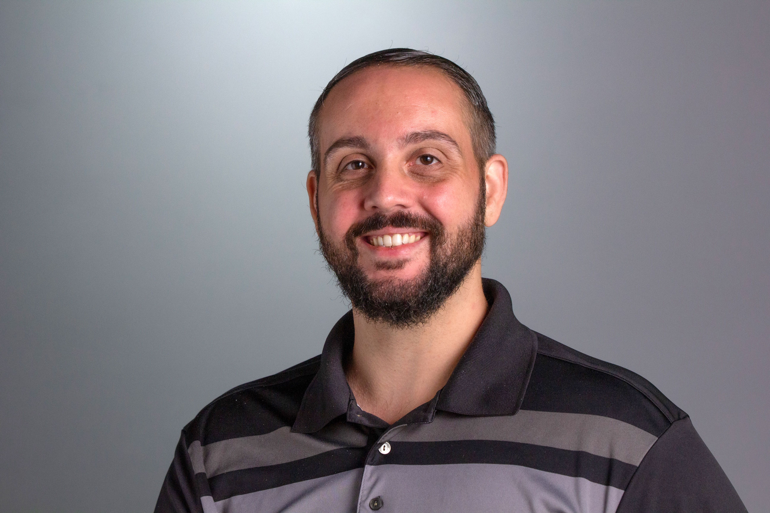 Headshot of Pat, who is sitting against a gray background smiling at the camera wearing a shirt with bold stripes. 