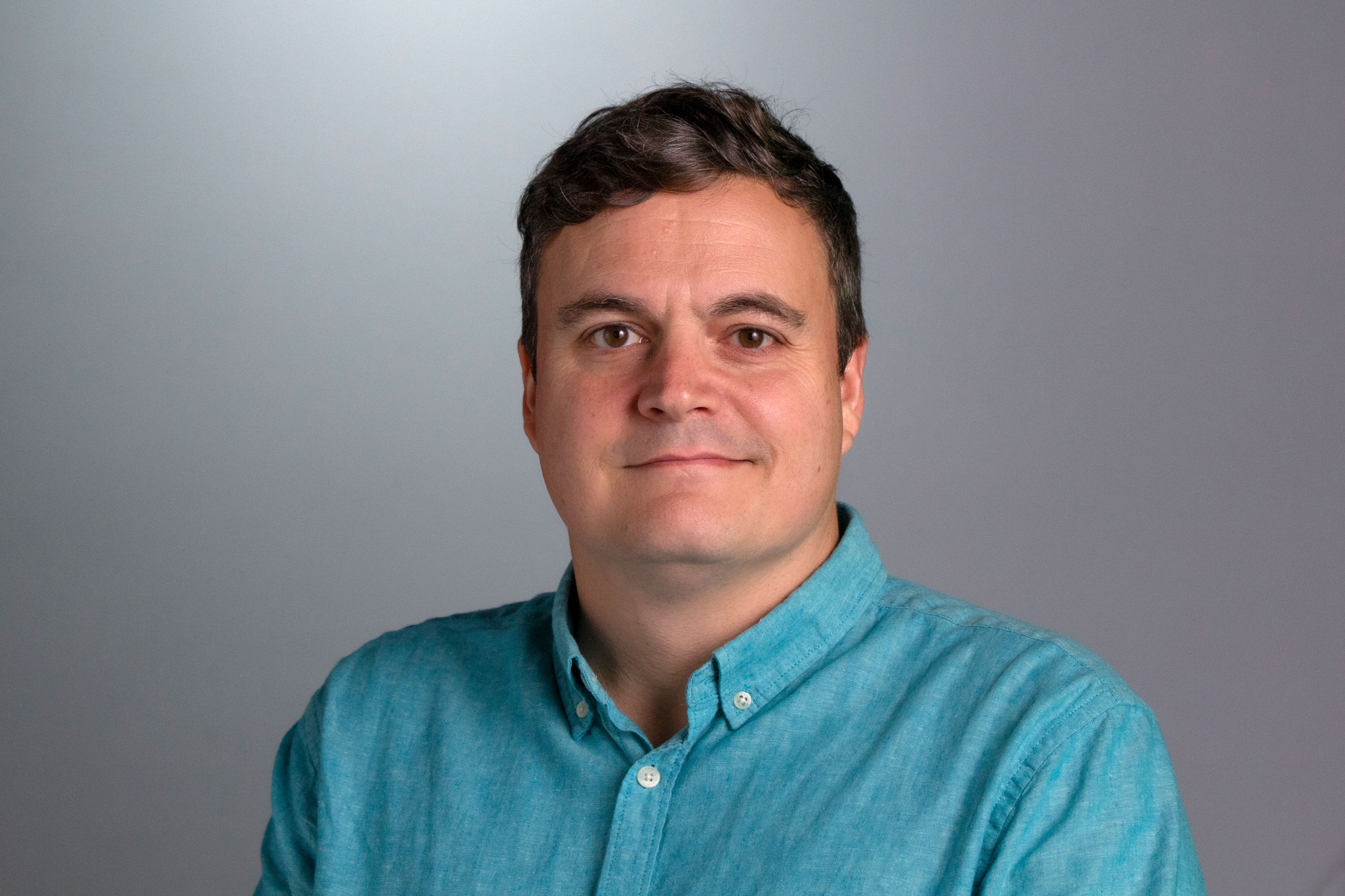 Headshot of Paul who is sitting against a gray background wearing a light blue button down shirt. 